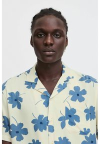Cămașă cu mâneci scurte, florală, de culoare galben deschis, cu modele de hibiscus albastre. Prezintă un guler deschis și un buzunar pe piept, fiind fabricată dintr-un material ușor.
