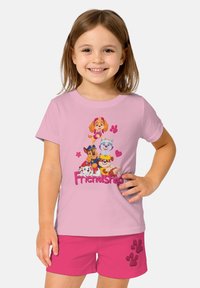 Ragazzina sorridente che indossa una maglietta rosa e pantaloni corti con cuccioli di cartone e la scritta "Amicizia" sulla maglietta, insieme a impronte di zampa sui pantaloni.