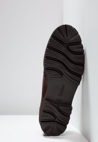 Semelle d'une chaussure Gabor marron foncé avec des rainures profondes, positionnée verticalement contre un mur d'angle blanc et le sol.