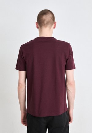 Personne aux cheveux courts portant un t-shirt bordeaux uni à manches courtes et un pantalon noir, vue de dos sur un fond uni.