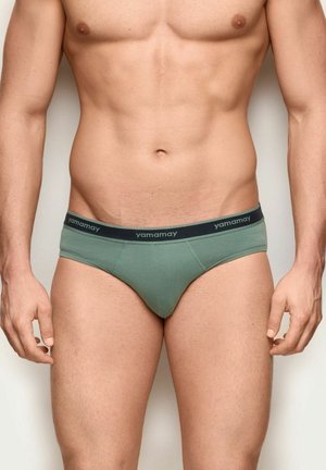 Fit männlicher Oberkörper in hellgrünen Slips mit dunkelfarbigem Bund, der mit "yamamay" beschriftet ist, vor einem schlichten neutralen Hintergrund stehend.