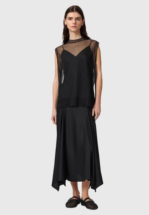 Ženska nosi črno prozorno brezrokavnik čez črno obleko na tanke naramnice, kombinirano s črnimi tkaničastimi ravnimi čevlji, stoji pred enobarvnim ozadjem.