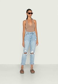 Top sem mangas castanho, jeans rasgados azul claro e sandálias de onça castanhas. Acessórios incluem óculos de sol escuros. De pé sobre um fundo liso.