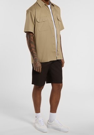 Persona che indossa una camicia beige a maniche corte con bottoni sopra una t-shirt bianca, pantaloncini neri, calzini bianchi e sneakers bianche, in piedi davanti a uno sfondo semplice.