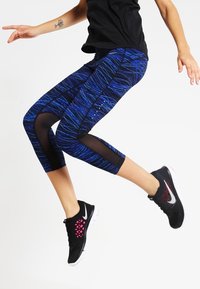 Leggings capri a fantasia blu con pannelli in rete, abbinati a scarpe da ginnastica nere e rosa. Texture liscia e vestibilità elastica per un uso attivo.