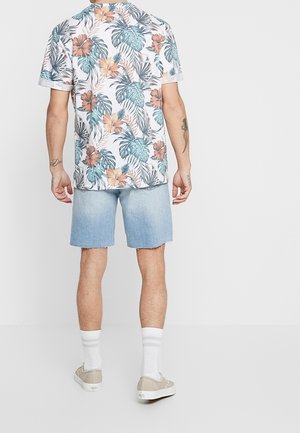 Weißes Kurzarmhemd mit mehrfarbigem tropischem Blumenmuster, kombiniert mit hellblauen Jeansshorts und beigen Sneakers, akzentuiert durch weiße Socken.