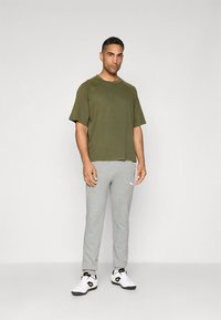 Olívazöld rövid ujjú póló, bő fazon, világosszürke melegítőnadrággal, melyen fehér logó található, fekete-fehér sportcipővel párosítva.