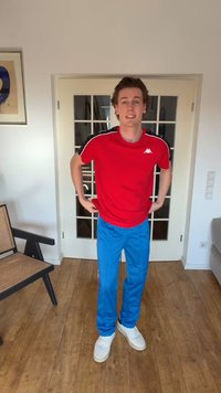 Νέος άνδρας στέκεται μέσα, φοράει κόκκινο αθλητικό μπλουζάκι, μπλε παντελόνια και λευκά αθλητικά παπούτσια μπροστά από διπλές γυάλινες πόρτες.