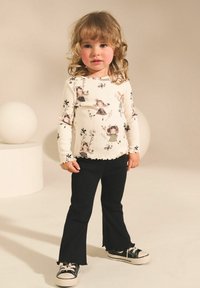 Langärmliges cremefarbenes Shirt mit floralen Illustrationen, kombiniert mit schwarzen Schlaghosen und schwarzen Sneakers mit weißen Sohlen, vor einem hellen Hintergrund stehend.