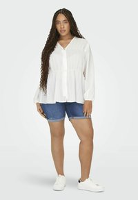 Balta ilgaa rankovėmis, mygtukais užsegama bliuska su klostėmis ir laisvu siluetu, derinama su tamsiomis džinsinėmis šortais ir baltais sportiniais bateliais.