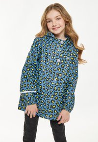 Giacca impermeabile blu con stampa leopardata nera e verde, cappuccio, chiusura a scatto e polsini elasticizzati, indossata sopra pantaloni neri.