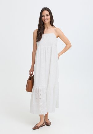 Femme en robe midi blanche sans manches en broderie anglaise, tenant un sac à main marron et portant des chaussures plates marron sur un fond blanc uni.