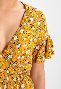 Gul blommig klänning med djup V-ringning, volangärmar och smal midja. Har små rosa och vita blommor på en strukturerad bakgrund.