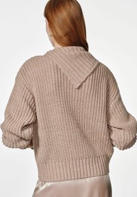 Sweter dzianinowy w jasnym beżu z kapturem, textured materiał, luźny krój oraz ściągacze na mankietach i u dołu. Posiada swobodny, luźny design na plecach.