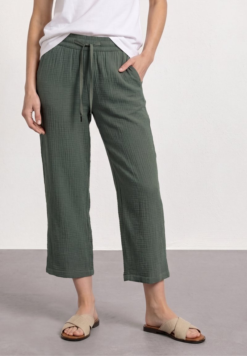 Femme portant un pantalon à cordon vert olive texturé et des sandales plates beiges à brides croisées, debout contre un mur blanc uni.