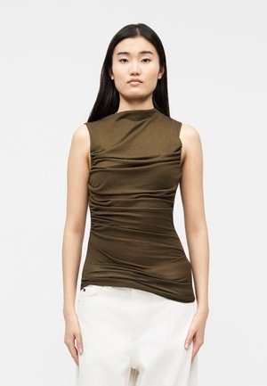 Mladá žena s dlhými tmavými vlasmi, oblečená v bezrukávovom olivovo zelenom vrúbkovanom topu a bielych nohaviciach s vysokým pásom, stojaca pred jednoduchým pozadím.