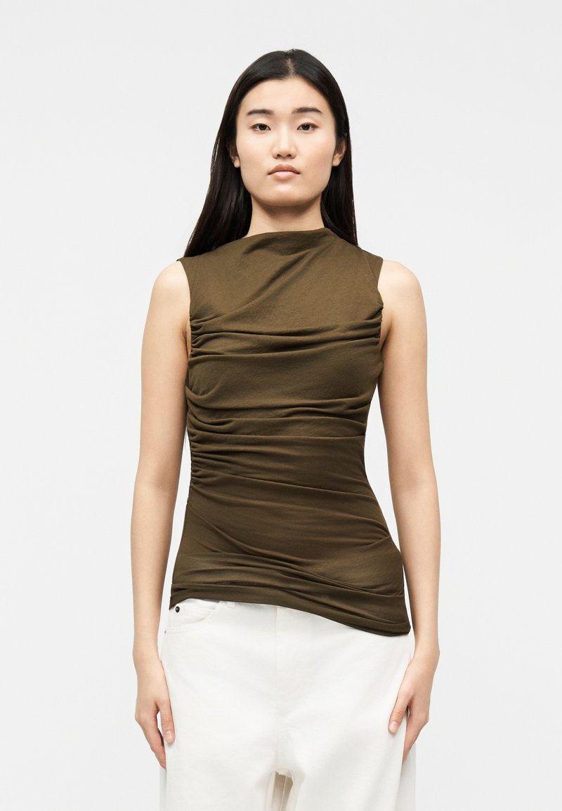 Fiatal nő hosszú sötét hajjal, ujjatlan, olívazöld, ráncolt felsőt és magas derekú fehér nadrágot viselve, egyszerű háttér előtt állva.