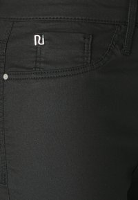 Svarta jeans med en slät, strukturerad tygkvalitet, med en silverfärgad logotyp på bakfickan och detaljerad sömnad längs sömmarna.