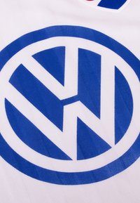 Witte stof met een blauw Volkswagen-logo, met gedurfde geometrische vormen en een gestructureerd oppervlak. Horizontale strepen zijn zichtbaar in het materiaal.