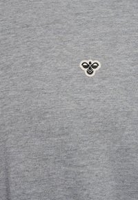 Petit logo d'abeille noir et blanc brodé sur le côté supérieur gauche d'un fond en tissu gris clair.