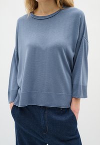 Svetlo modra pletena pulover s okroglo izrezom, ohlapen kroj in tričetrtinski rokavi. Rebrast rob dodaja teksturo. V kombinaciji s temno jeans hlačami.