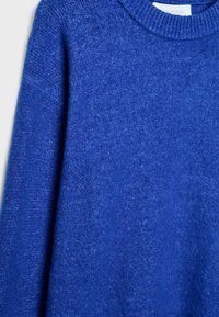 Modro pleten pulover z okrolim izrezom. Mehka tekstura in rebrasti detajli na manšetah in robu. Brez vidnih vzorcev ali poudarkov.
