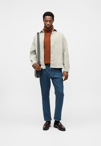 Veste gris clair, col roulé rouille, jeans bleus, chaussures noires. Le modèle tient un sac noir, doté d'une texture lisse et de détails à boutons.