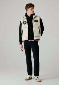 Mladý muž ve smetanové fleecové vestě přes černou mikinu, tmavých džínách a bílých teniskách, stojí a dívá se do strany na jednolitém pozadí.