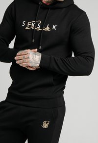 Felpa nera con lettering dorato, dotata di cappuccio con lacci e polsini a coste. Abbinata a pantaloni neri con logo dorato.