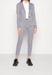 Traje con estampado que presenta un diseño geométrico en azul, rojo y blanco. La chaqueta tiene hombros estructurados y los pantalones son de corte ajustado. Calzado negro.