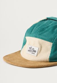 Roheline ja kreemja värviga kübar, millel on pruun ääreosa ja esipaneelil tikitud silt. Tekstuur on ribaline, mis lisab visuaalset huvi.