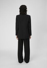 Blazer noir sur mesure à coupe droite, manches longues et design minimaliste, associé à un pantalon noir à jambes larges.