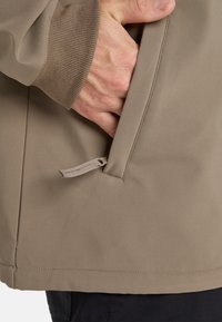 Smeđa softshell jakna s rebrastim manžetama, koja sadrži bočnu džep i detalj sa zatezanjem na džepu, dizajnirana za aktivnosti na otvorenom.