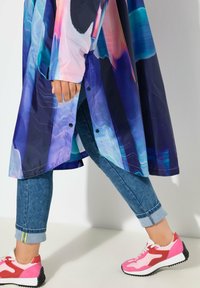 Bunte, glänzende lange Jacke mit blauen, pinken und lila abstrakten Mustern; kombiniert mit hochgekrempelten Jeans und pinken Sportschuhen.