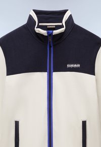 Giacca in pile blu navy e crema con colletto alto, zip frontale, tasche laterali e un accento blu sulla zip. Presenta un logo sul lato sinistro superiore.