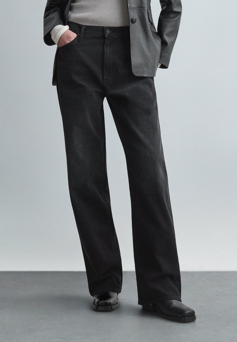 Oseba, oblečena v ohlapne črne kavbojke, črne usnjene čevlje, sivi blazer in belo dolgo majico z dolgimi rokavi, z rokami v žepih.