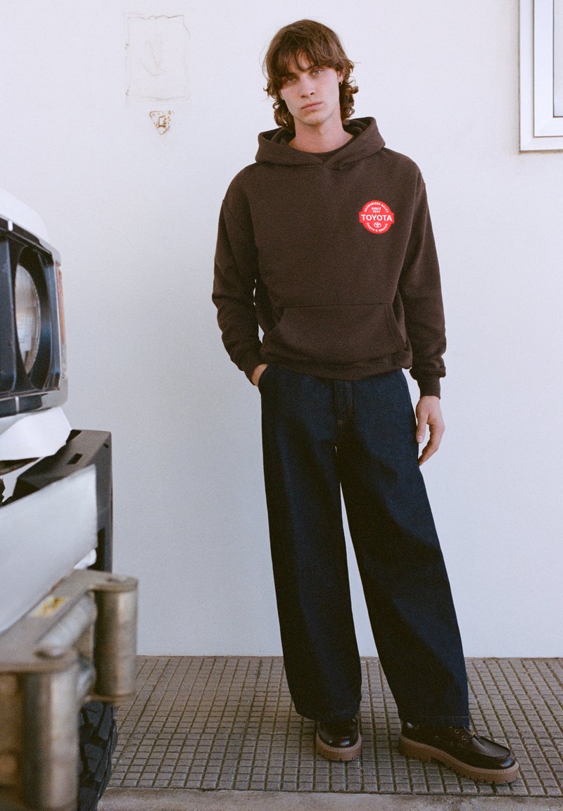 Hnedá mikina s kapucňou s červeným logom Toyota, tmavomodré široké nohavice a čierne topánky s svetlohnedými podrážkami. Jednoduché pozadie s vozidlom.