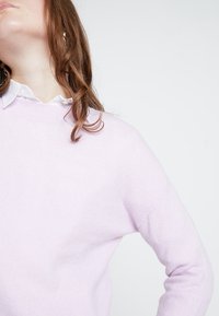 Pull lavande claire en matériau doux, avec un col rond et des manches longues, porté par-dessus une chemise blanche à col.