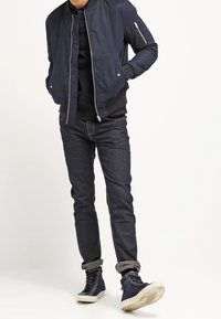 Marinblå bomberjacka med en zipficka, kombinerad med mörk denimjeans och marinblå högskaftade sneakers med vita sulor. Skräddarsydd passform, slät textur.