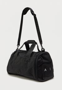 Μαύρο νάιλον σακίδιο με στρογγυλό σχήμα, διαθέτοντας διπλές λαβές κορυφής και αποσπώμενο ιμάντα ώμου, λογότυπο ανάγλυφο και φερμουάρ κλεισίματος.