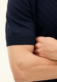 Mörkblå stickad kortärmad tröja med en strukturell diamantmönster och ribbad mudd, som visar en del av en arm som korsar en hand.