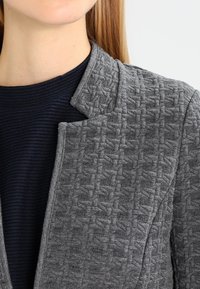 Szary, teksturowany żakiet z plecionym wzorem, bez kołnierza. Nosi się go na ciemnoniebieskiej, prążkowanej koszuli.