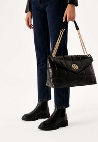 Black quilted leather handbag with a gold hardware logo, featuring dual chain straps and a flap closure. Worn with dark jeans and ankle boots.