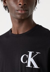Svart bomulls T-skjorte med en hvit brodert «cK»-logo på venstre bryst. Stoffet ser glatt ut med en klassisk rund hals.
