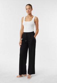 White tank top paired with loose black trousers featuring pockets and drawstring waist. Model wears white strappy sandals.