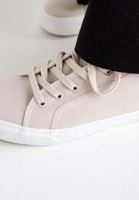 Beige vászontornacipő fehér gumitalppal, kerek orral és lapos fűzővel. Finom varrásokkal és sima felső résszel rendelkezik.