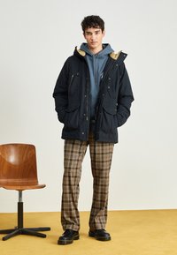 Navy parka jacket with a beige lining, blue hoodie, plaid brown pants, and black shoes. Features multiple pockets and a relaxed fit.