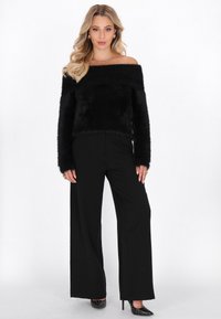 Maglione nero off-shoulder in tessuto peloso abbinato a pantaloni neri a gamba larga. L'outfit presenta una trama morbida e una vestibilità rilassata, completato da tacchi con borchie.