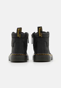 Botas negras hasta el tobillo con un acabado texturizado, cremalleras laterales, acentos amarillos y una suela gruesa con un pespunte amarillo en contraste.
