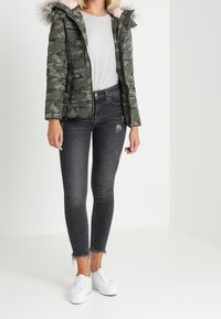 Polstret camouflagejakke med hætte i pelskant, parret med tætsiddende sorte jeans og hvide sneakers. Glatte teksturer og moderne snit.